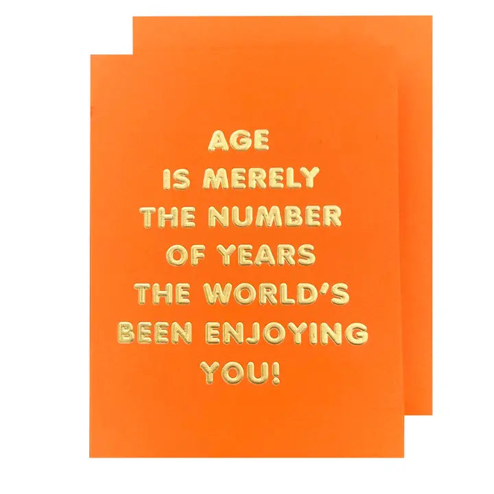 An orange greeting card with the text 'Age is merely the number of years THE WORLD'S BEEN ENJOYING YOU!' printed in gold. The card also has a smaller text that reads 'Blank Inside'.