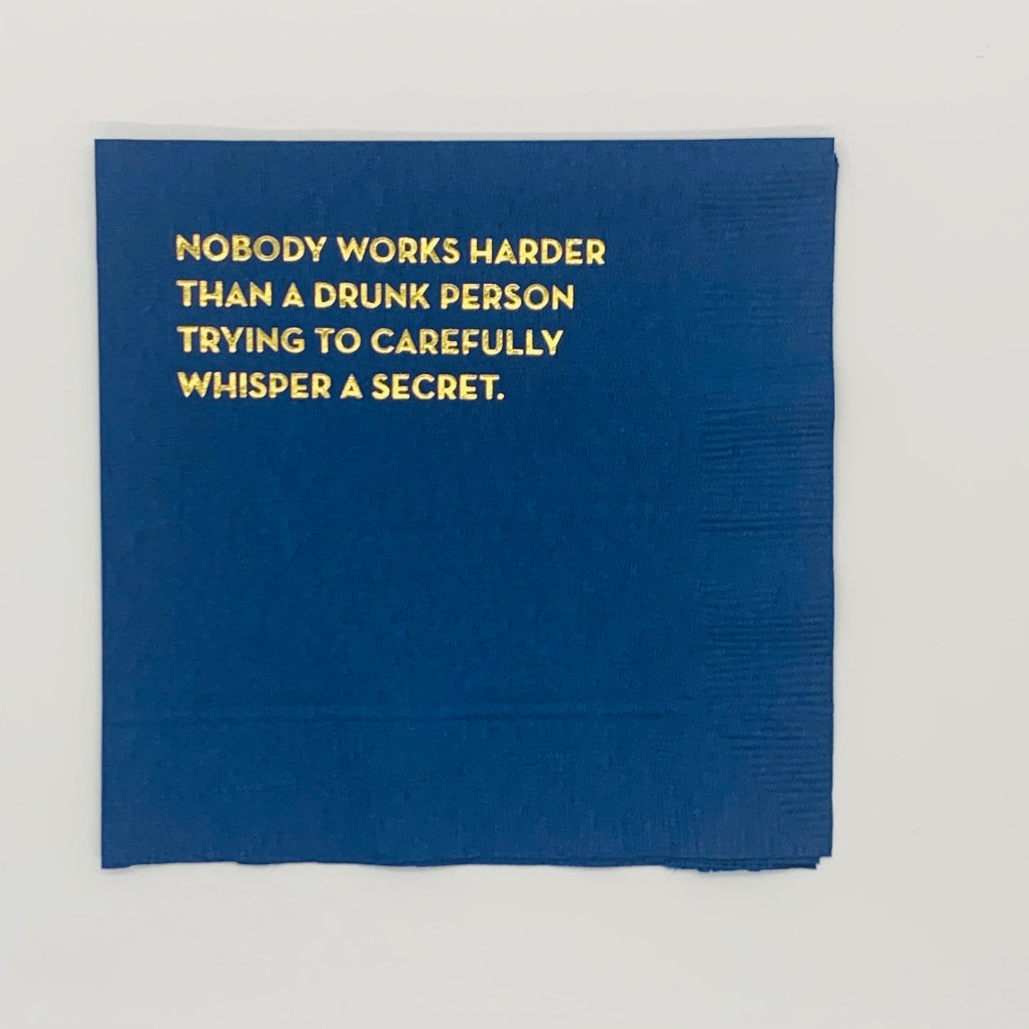 A navy blue cocktail napkin with gold foil print text 'Nobody works harder than a drunk person trying to carefully whisper a secret.'.