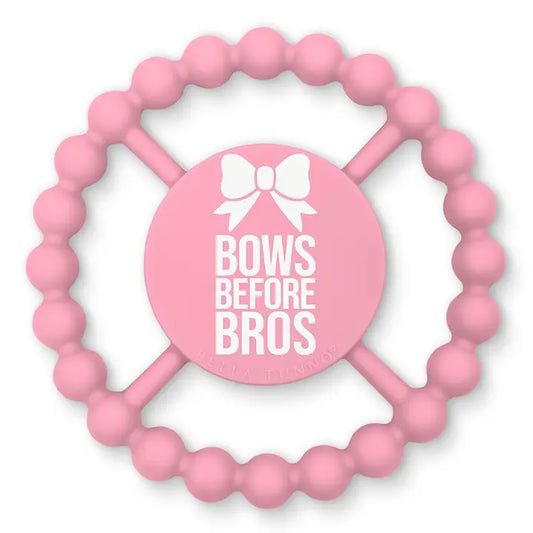 Pink silicone baby teether with a circular beaded ring and a central round pad featuring the text 'BOWS BEFORE BROS' and a white bow graphic by Bella tunno