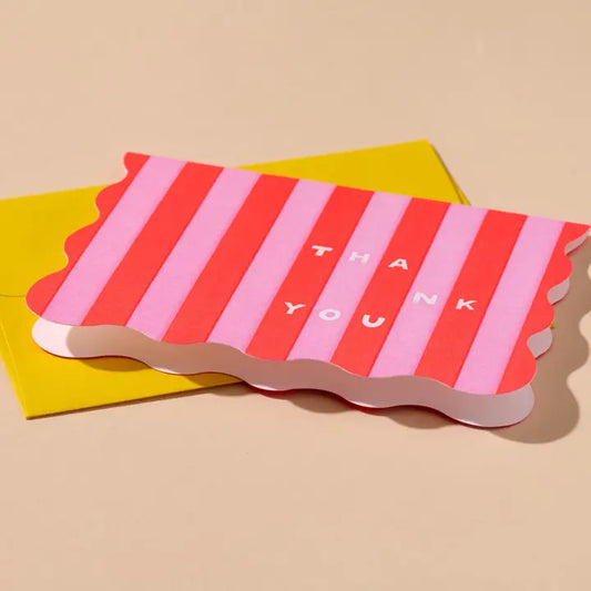 A scalloped thank-you card with alternating pink and red stripes and the words 'THANK YOU' printed in white, layered over a bright yellow envelope.