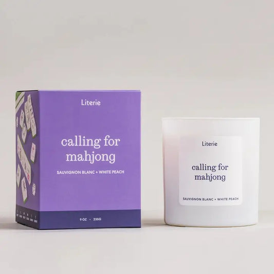 Literie candle in a white glass tumbler with white label that reads "calling for Mahjong" in black lettering and purple packaging next to it with a graphic of mahjong tiles on the side