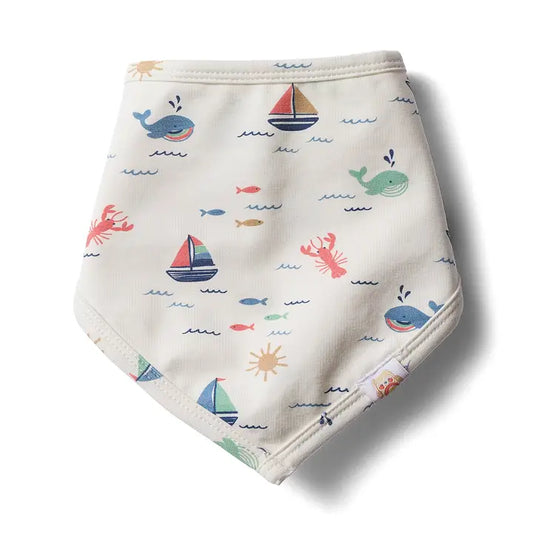 An off-white cotton baby bib with a colorful nautical print featuring blue whales, blue and red sailboats, red lobsters, fish, and yellow suns.