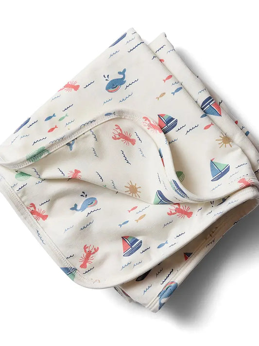 A folded baby blanket or swaddle with an all-over nautical print on a warm cream background. The print features blue whales, red lobsters , blue and red sailboats, and small yellow suns.