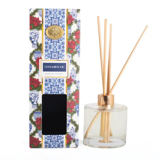 A reed diffuser bottle with natural wood reeds and a gold cap next to its patterned packaging box featuring blue and white ginger jars and red poinsettias.
