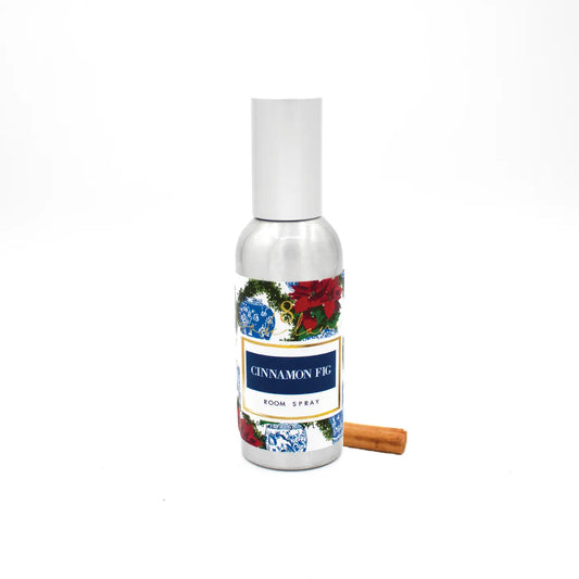 A silver room spray bottle labeled 'CINNAMON FIG ROOM SPRAY', with blue and white ginger jars and red poinsettia, shown on a white background with a cinnamon stick beside it.