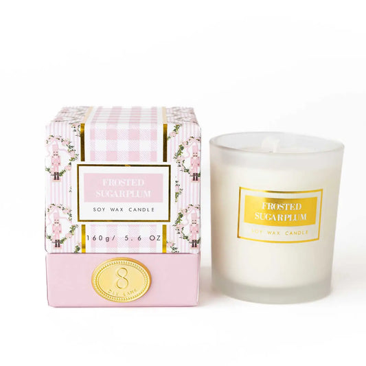 A frosted sugarplum scented soy wax candle in a clear glass jar next to its pink and white packaging box with pink florals, Nutcrackers, and gold accents, on a white background.