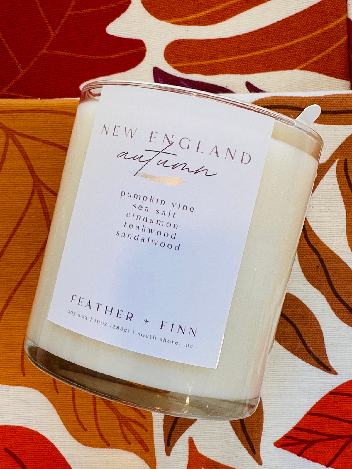 A New England Autumn scented candle by Feather + Finn in a clear glass jar, featuring a white label with gold and black text, resting on a patterned fabric background with orange, red, and cream leaf motifs.