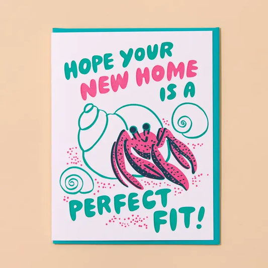 A greeting card with a white background and teal envelope, featuring a pink hermit crab illustration and the text 'HOPE YOUR NEW HOME IS A PERFECT FIT!' in teal and pink lettering.