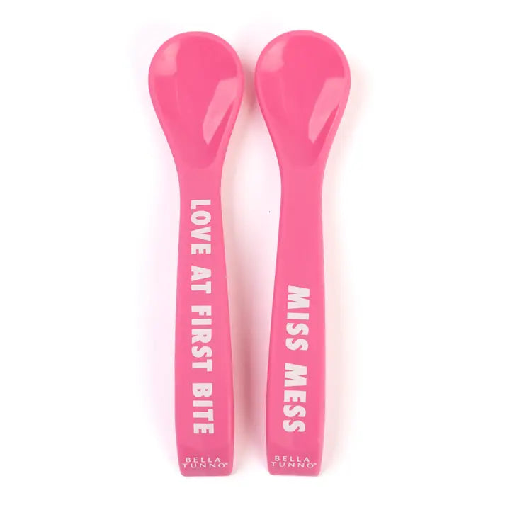 Two pink baby spoons with text on a white background that say "Love at First Bite" and "Miss Mess"