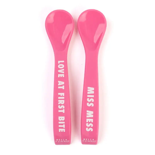 Two pink baby spoons with text on a white background that say "Love at First Bite" and "Miss Mess"