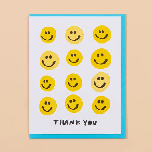 A greeting card with a white front, decorated with twelve yellow smiley faces in different expressions and the words 'THANK YOU' in black at the bottom. The card is shown with a bright blue envelope.