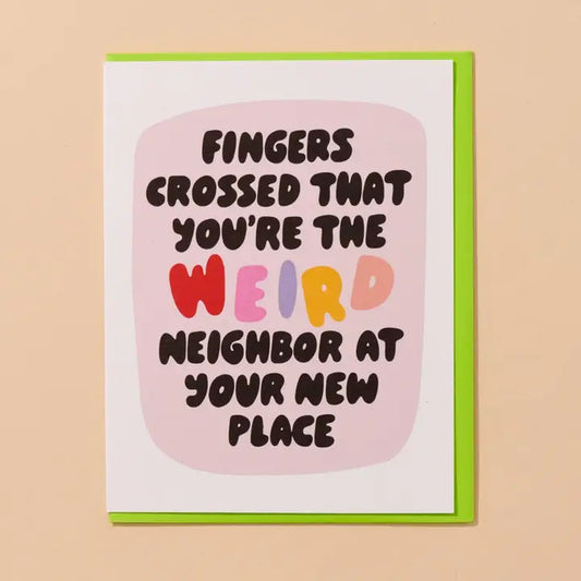 A greeting card with a bright green envelope, featuring the text 'FINGERS CROSSED THAT YOU'RE THE WEIRD NEIGHBOR AT YOUR NEW PLACE' in dark brown lettering over a pink rounded rectangle on a white card background.