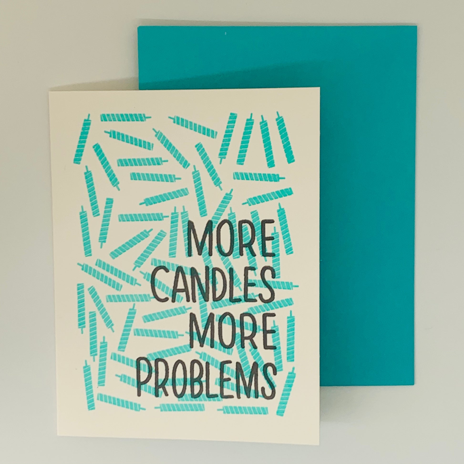 White Greeting Card with the text 'More Candles More Problems' with scattered teal candles.  Matching teal envelope
