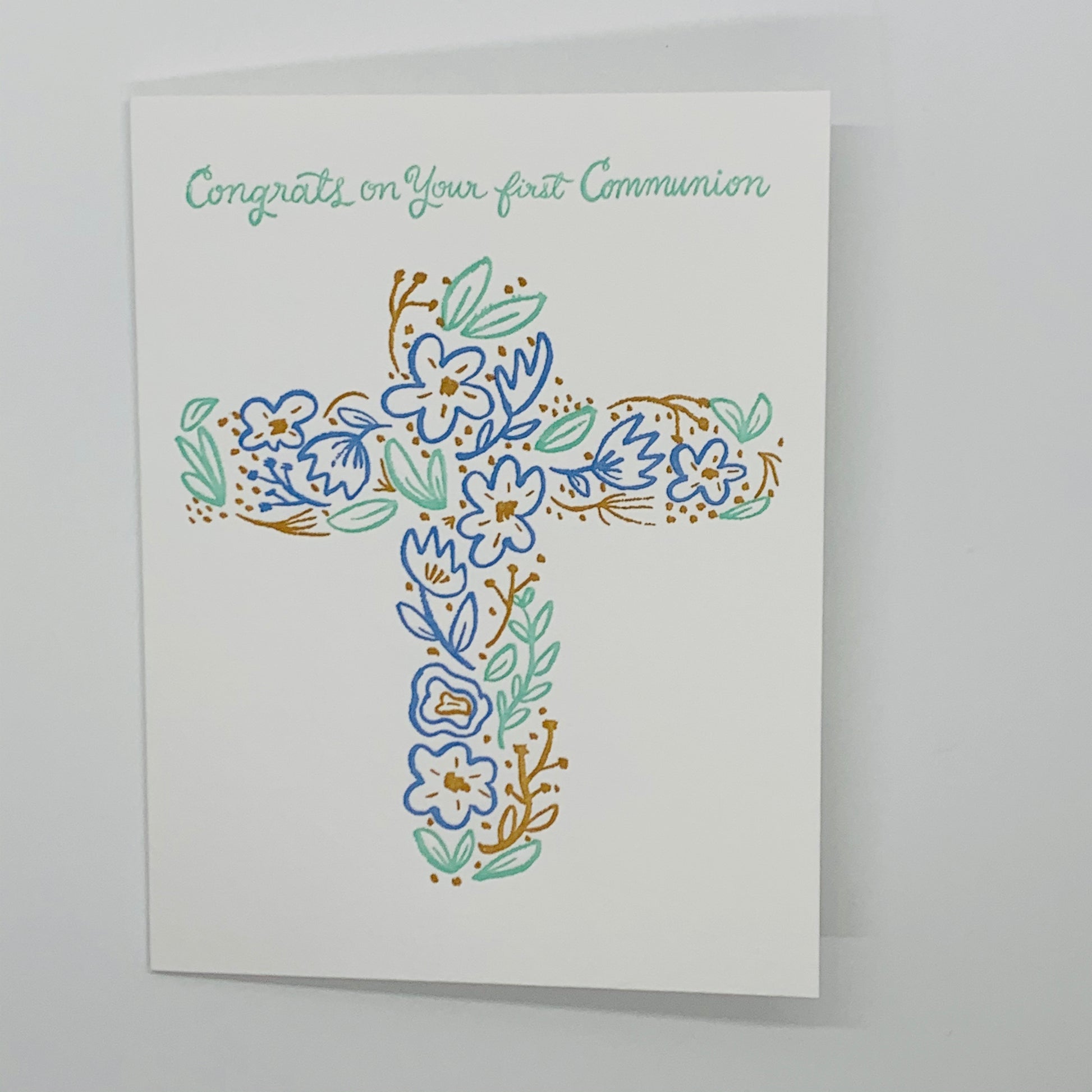 white card with congrats on your first communion in mint green.  a cross with blue, green, gold flowers